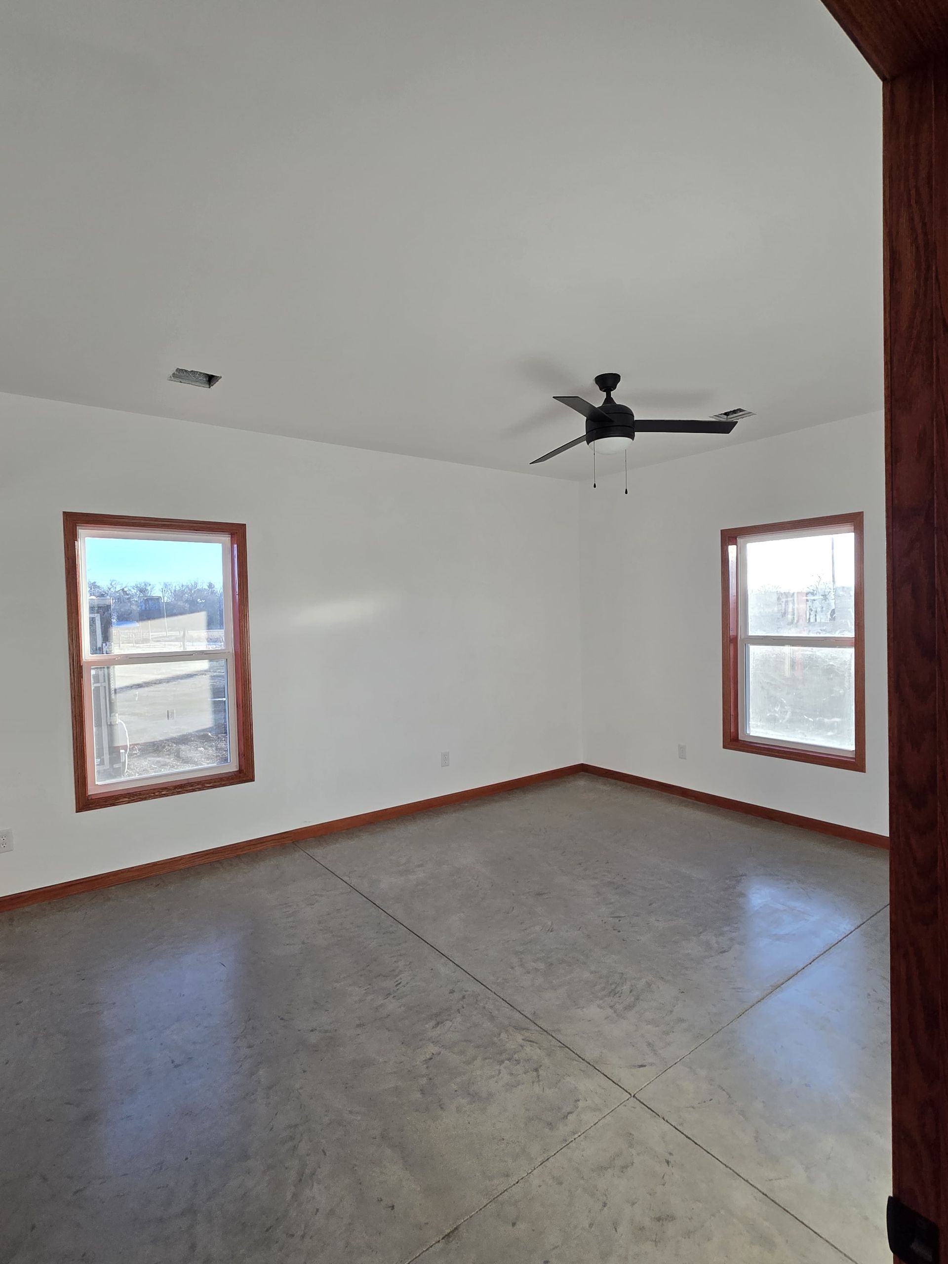 A room with white walls, two windows with wooden frames, a ceiling fan, and a gray concrete floor.