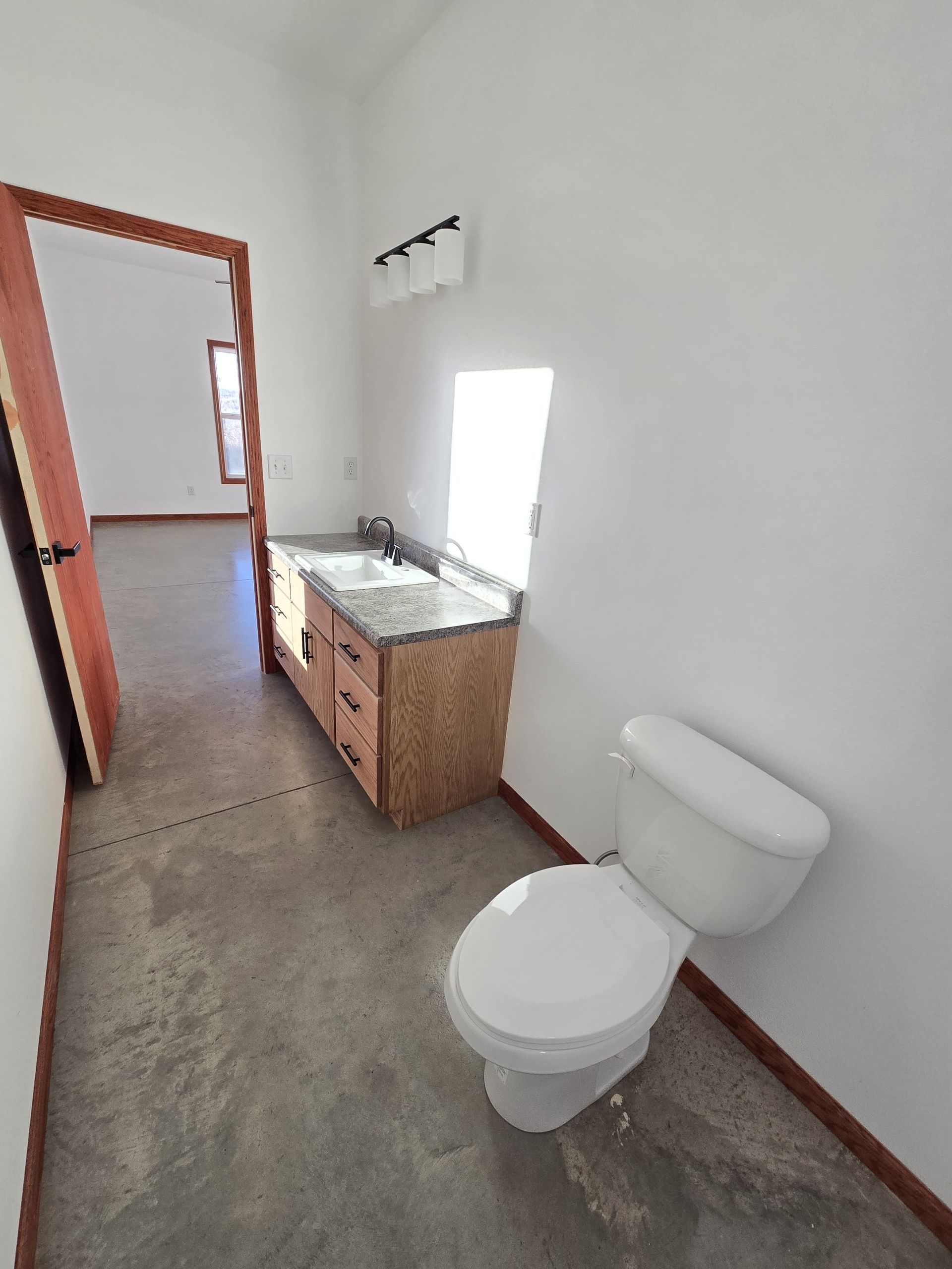 A bathroom with a white toilet, a wooden vanity with a stone countertop, a sink, and a light fixture against a white wall.