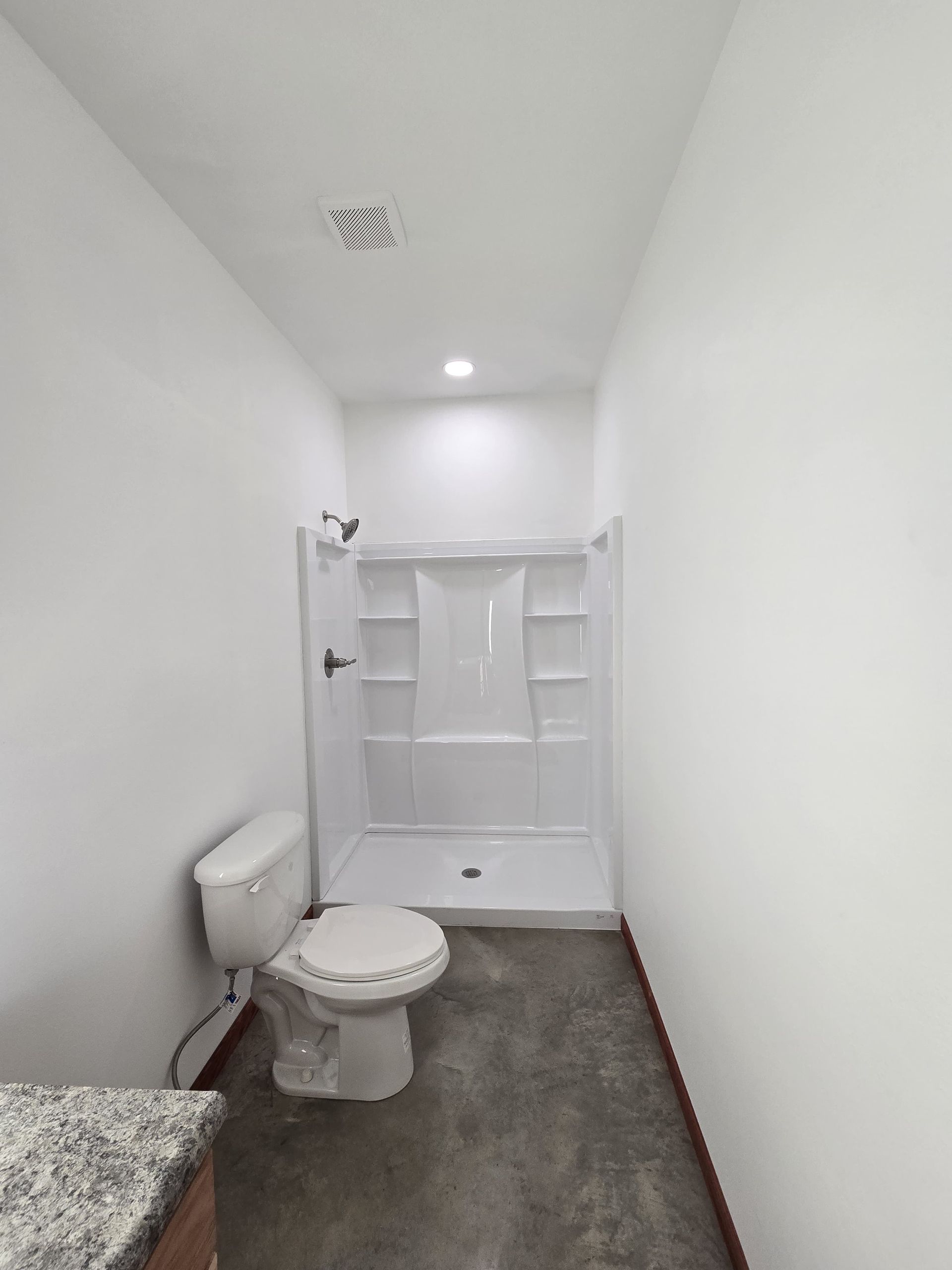 A bathroom with white walls, a gray floor, a white toilet, and a white shower stall with built-in shelves.