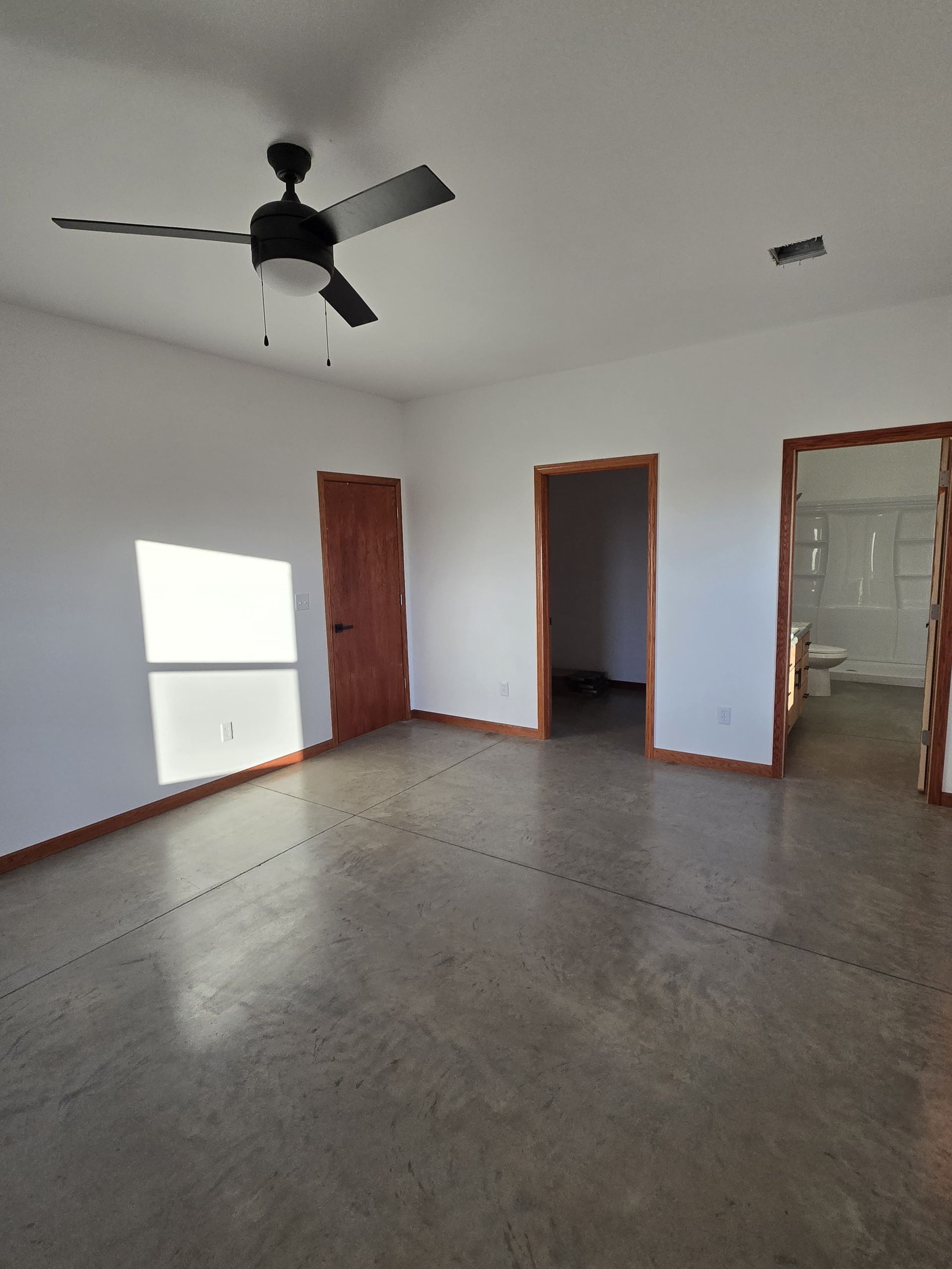 An empty bedroom with white walls, a black ceiling fan, gray concrete floors, and two doorways.