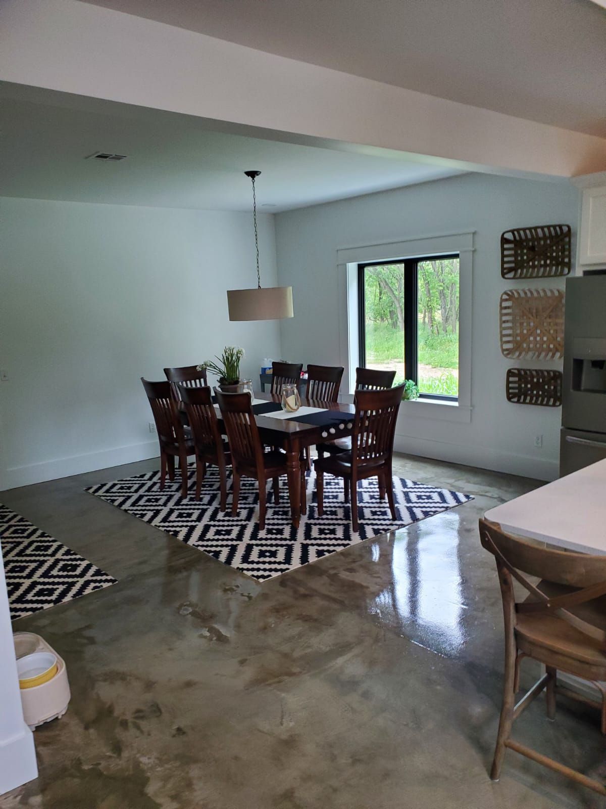 A dining area with a dark wood table and chairs on a geometric rug, set against polished concrete floors in a bright room.