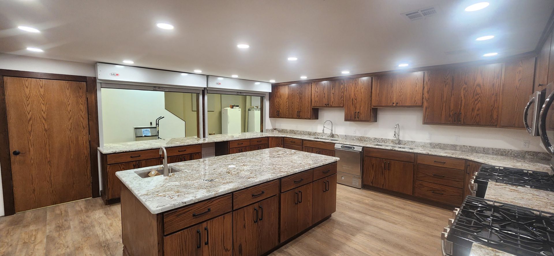 A wide-angle kitchen with wood cabinets, light granite countertops, an island with a sink, and recessed ceiling lighting.