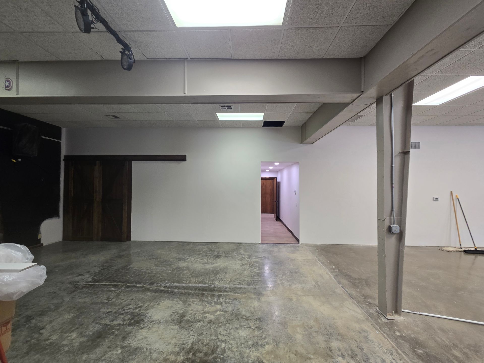 A wide, sparse commercial interior with concrete floors, white walls, dark wooden doors, and fluorescent lighting.