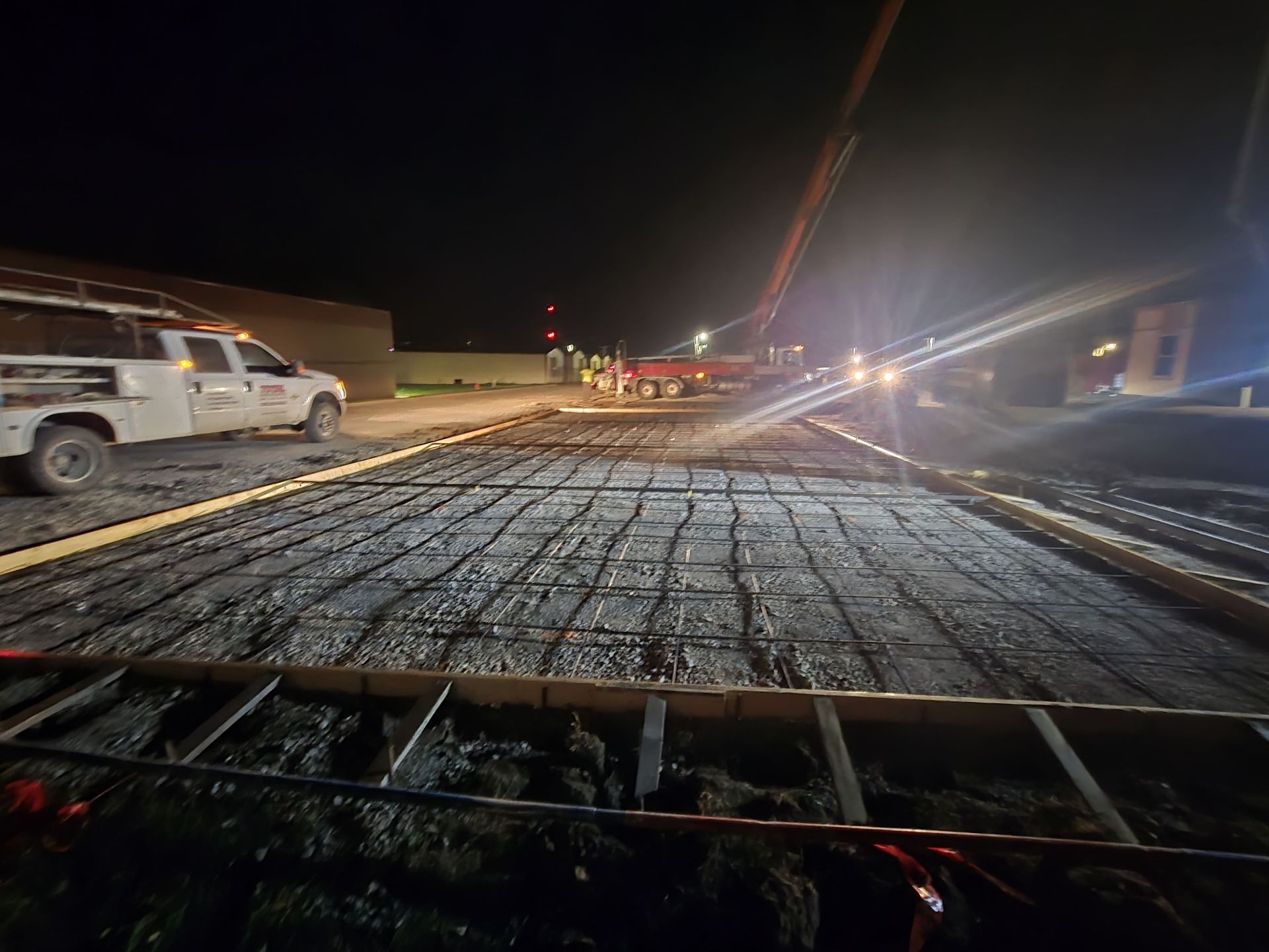 At night, a concrete pump truck pours cement over a metal rebar grid inside wooden construction forms.