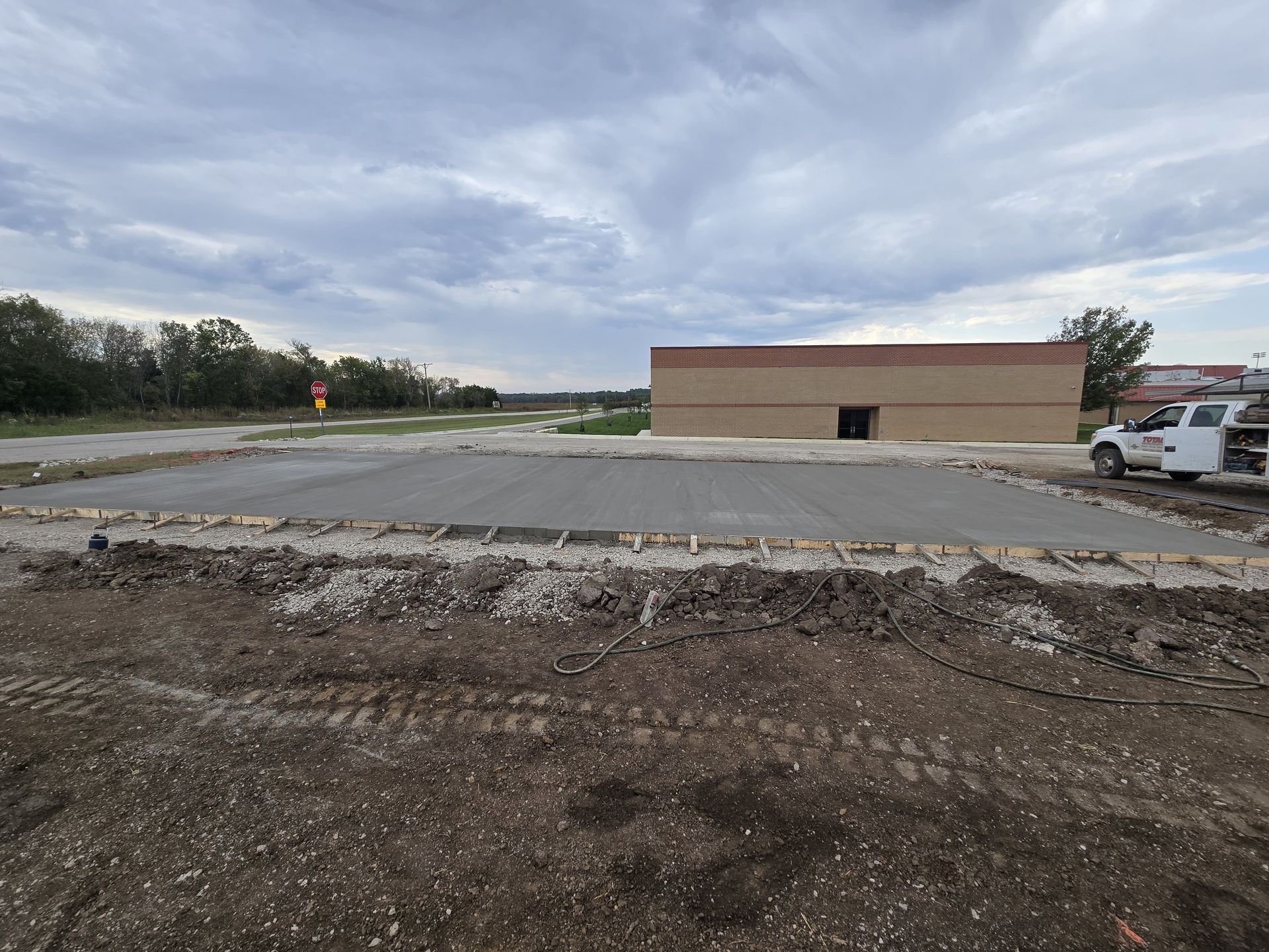 A freshly poured concrete slab sits in an open dirt lot, with a brown building and a parked truck in the background.
