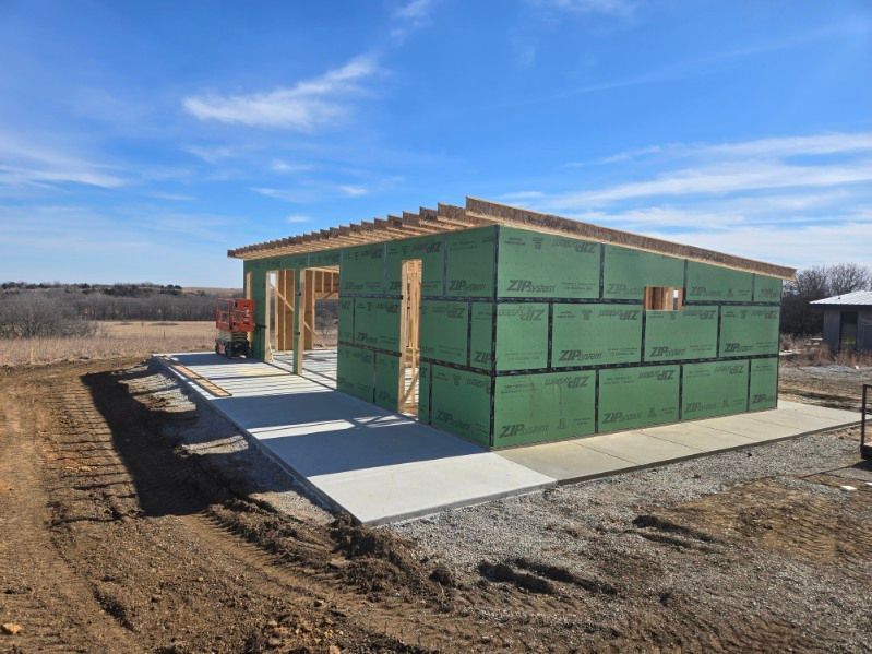 A partially constructed building on a concrete slab, framed with wood studs and sheathed in green panels under a blue sky.