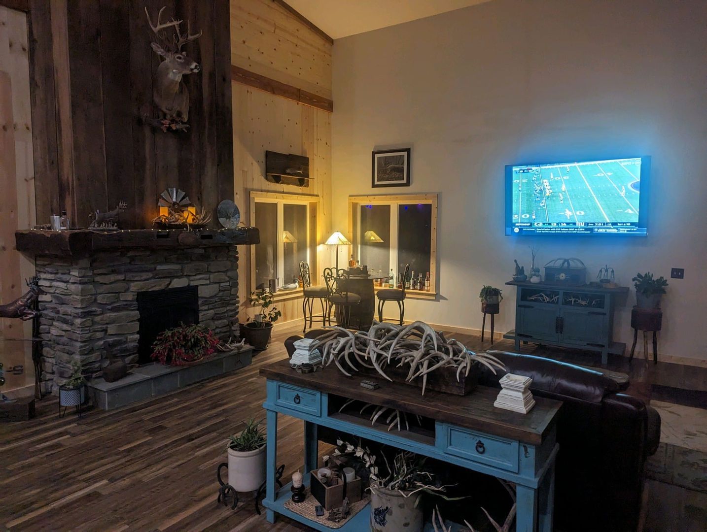A rustic living room with a stone fireplace, a wall-mounted TV, a blue console table with antlers, and a deer head mount.