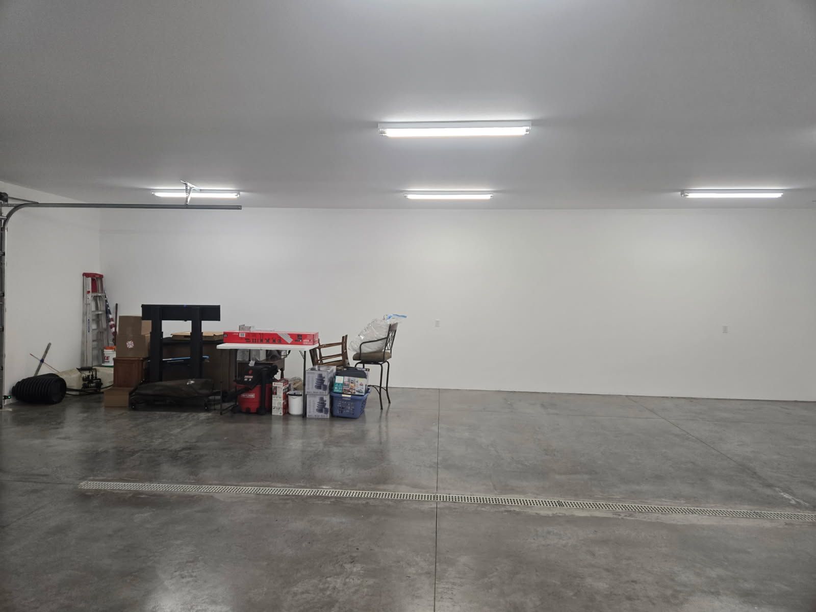 Empty indoor garage or warehouse with white walls, gray floor, and a small workbench with supplies on the left.