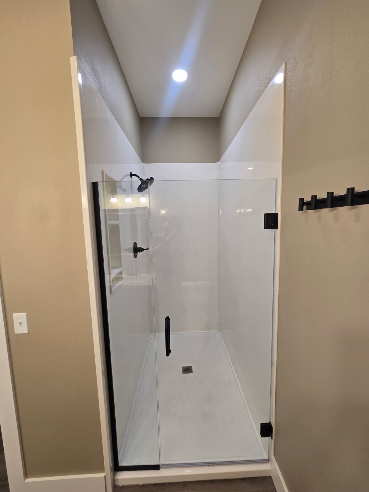 Glass shower enclosure in a beige bathroom with a high sloped ceiling and recessed light.