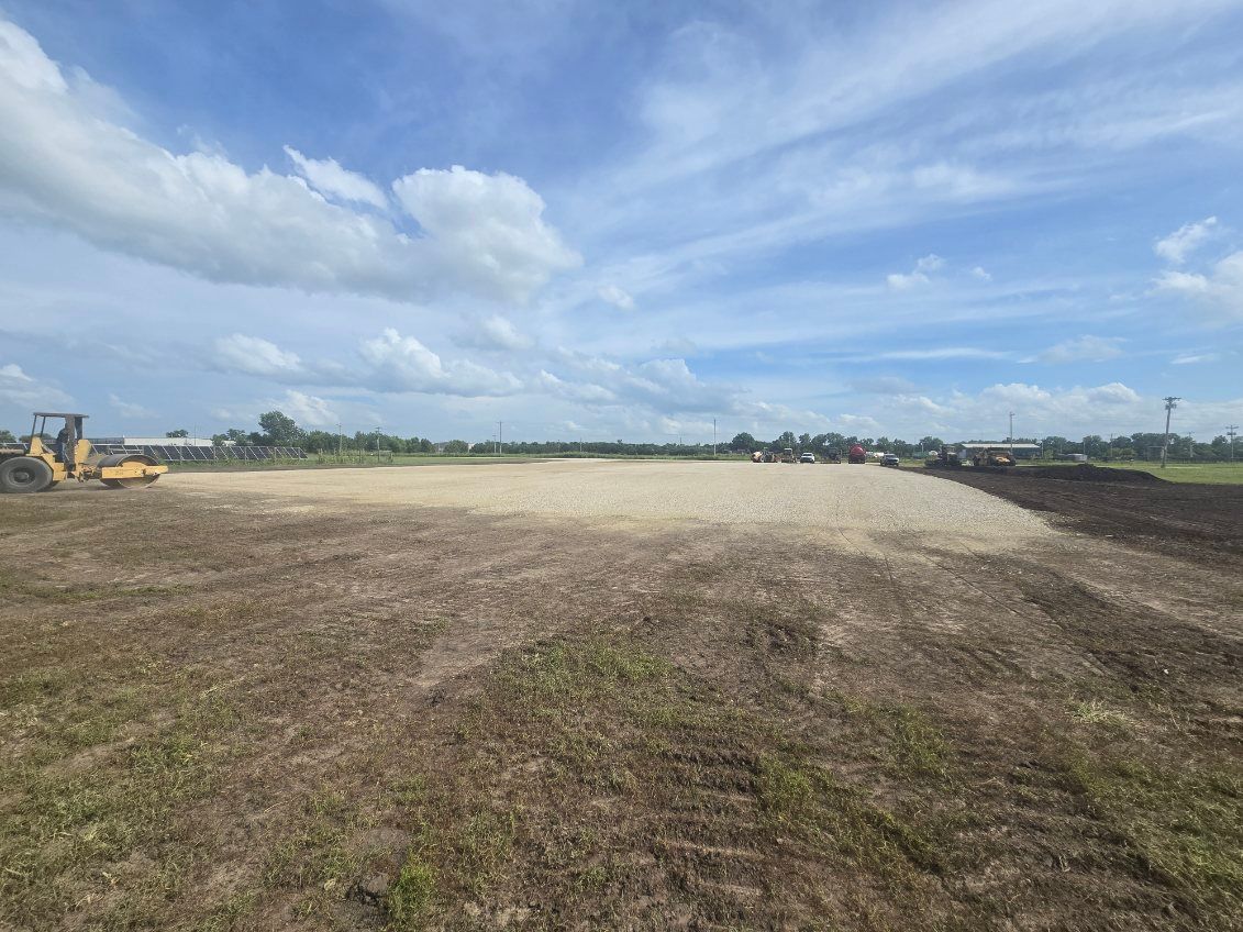 Wide dirt field under a blue sky with clouds, with construction equipment at the left edge.