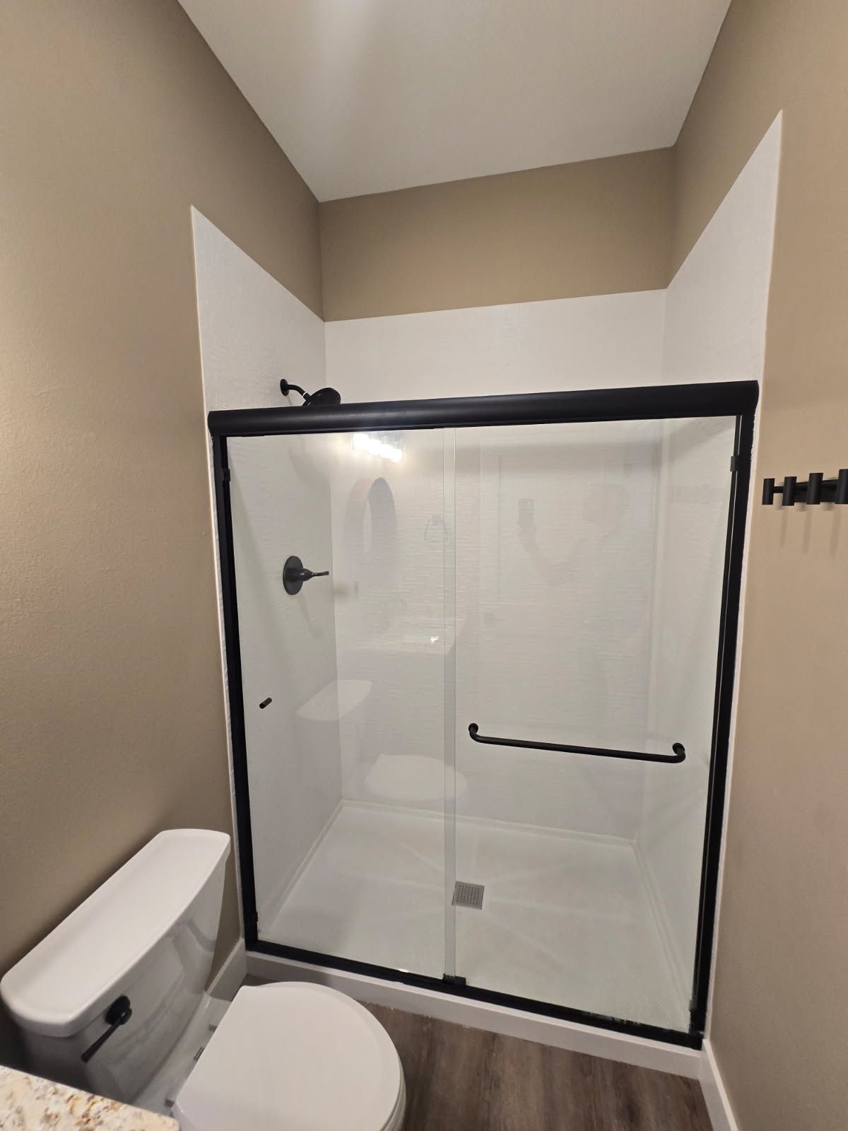 Small bathroom with white glass shower enclosure, toilet, beige walls, and black shower trim.