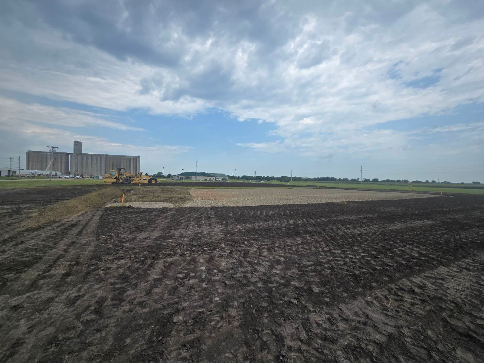 Freshly plowed field under a cloudy sky, with a row of buildings in the distance.