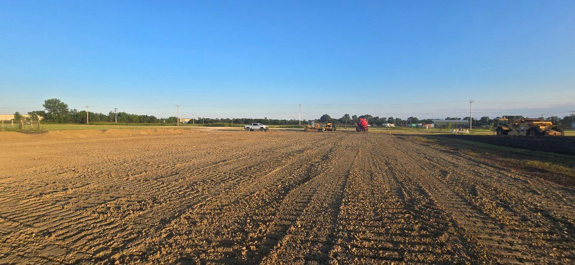 Wide plowed field under a clear blue sky at sunset, with distant farm equipment on the horizon