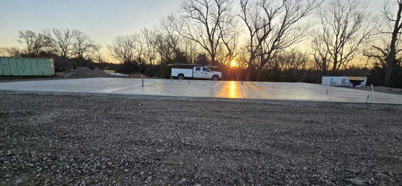 A newly poured concrete slab at a construction site at sunset, with a white utility truck parked near the trees.