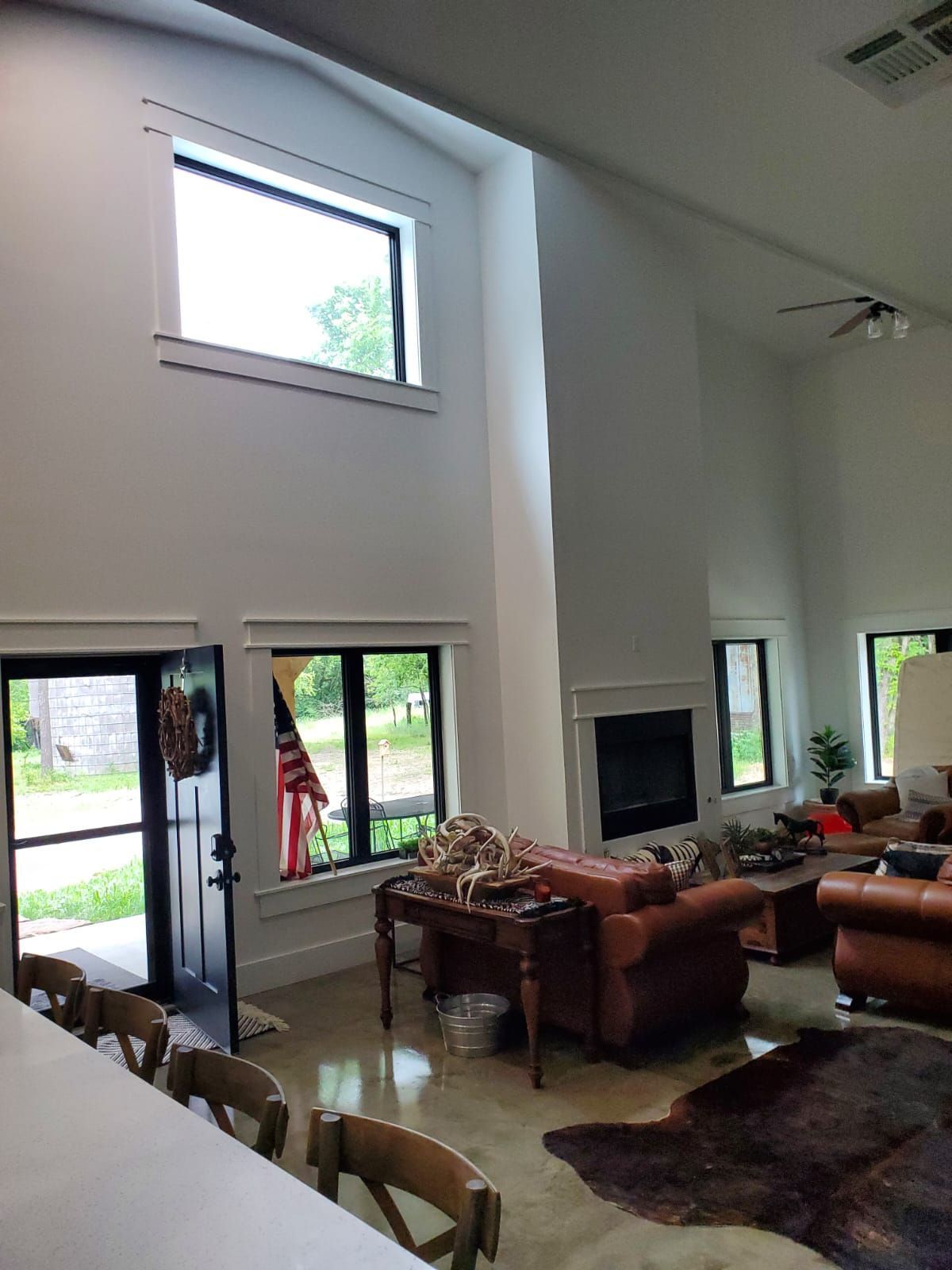 A high-ceilinged living room with white walls, large windows, dark trim, brown leather furniture, and concrete flooring.