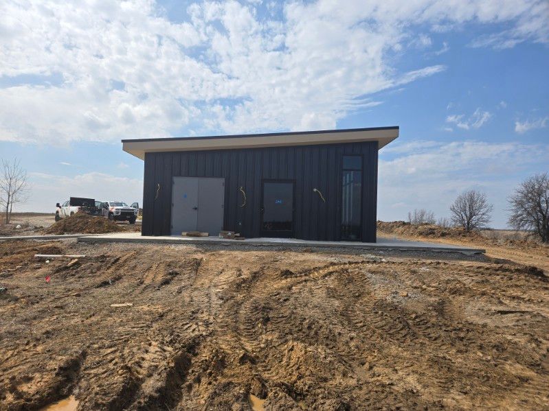 A modern black rectangular utility building with a flat roof, situated on a concrete slab in a dirt field under a blue sky.