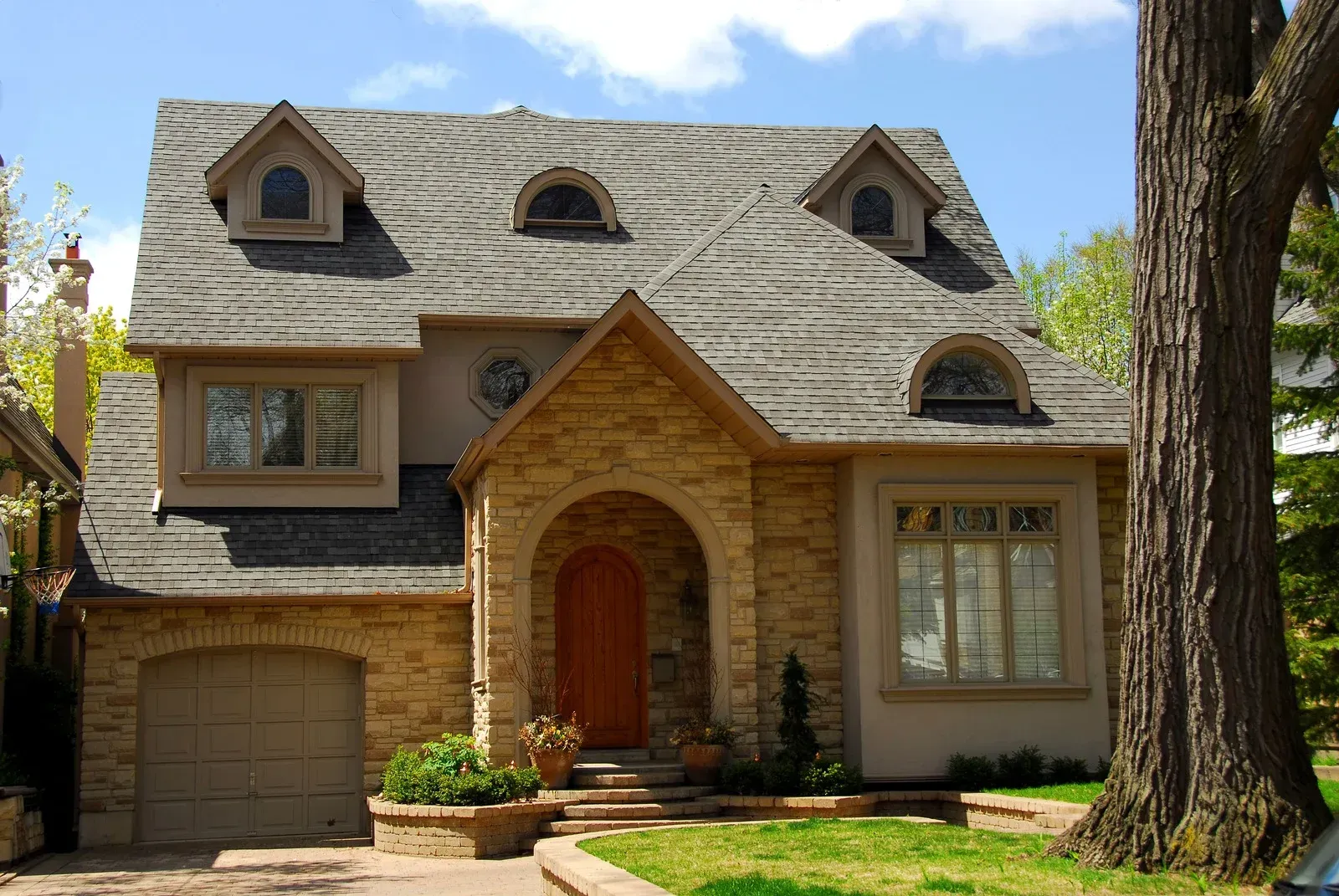 Stone house with arched doorway, dormer windows, and garage. Light brown stone and grey roof.
