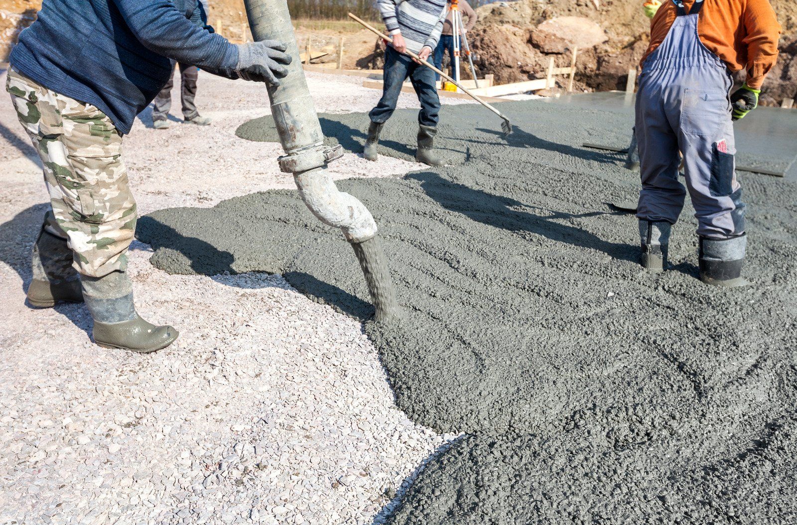 Workers pouring concrete onto a gravel base. A hose dispenses gray concrete; other workers use tools to spread it.