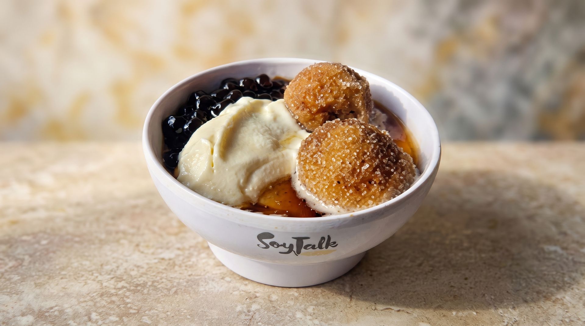 Small white bowl with ice cream and dessert balls, topped with dark sauce on a stone surface