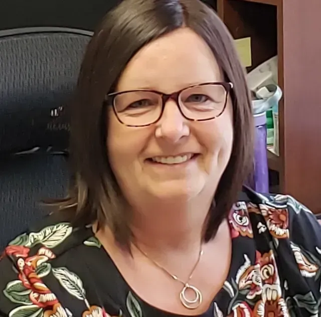 Woman with glasses smiling, wearing a floral top, in an office setting.