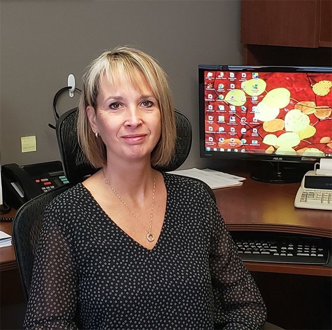 Woman in office setting, smiling at camera, sitting at a desk with a computer and phone.