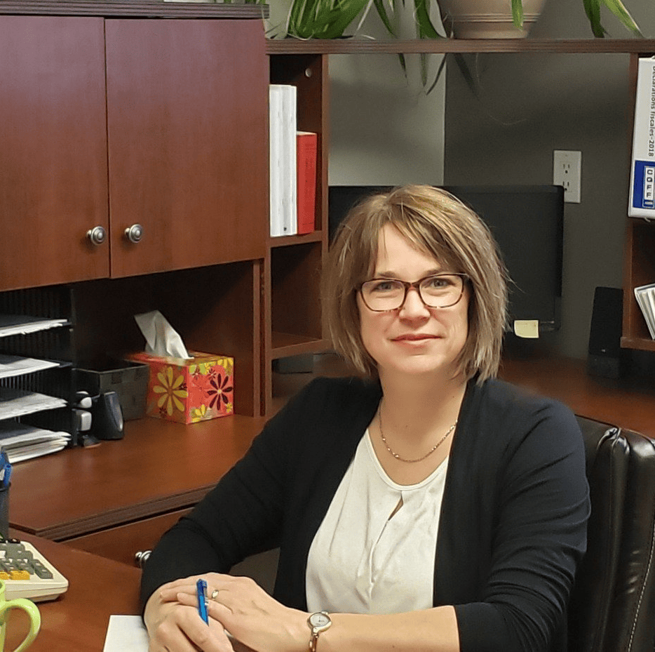 Woman in glasses at a desk in an office setting, smiling and holding a pen.