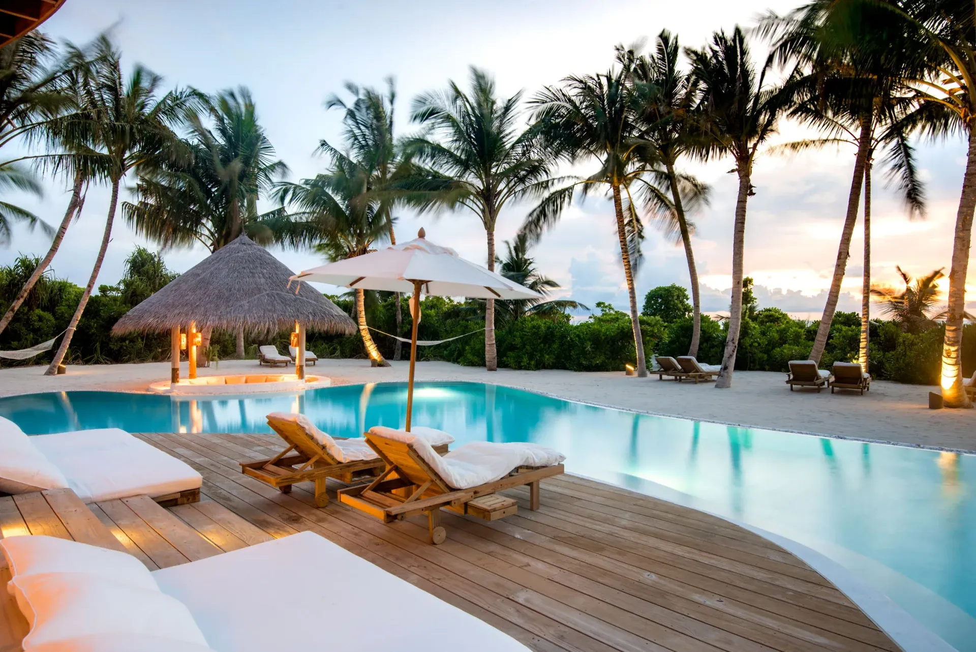Tropical resort pool with lounge chairs, white umbrellas, and palm trees at sunset.