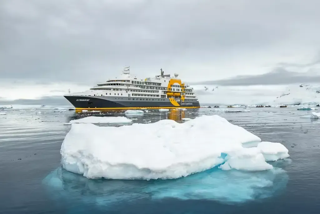 Icebreaker ship beside a large white iceberg in icy blue water under a cloudy sky