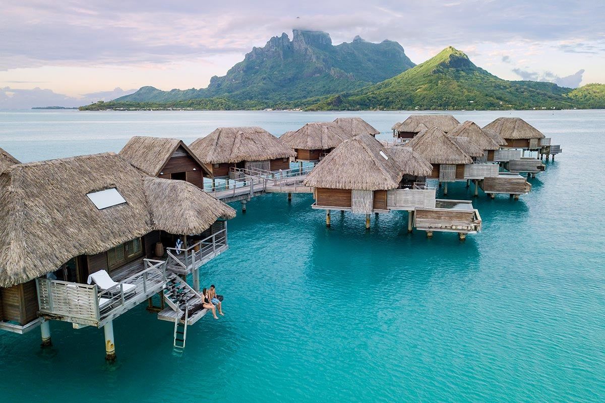 Overwater thatched bungalows on turquoise lagoon with lush green island in the distance