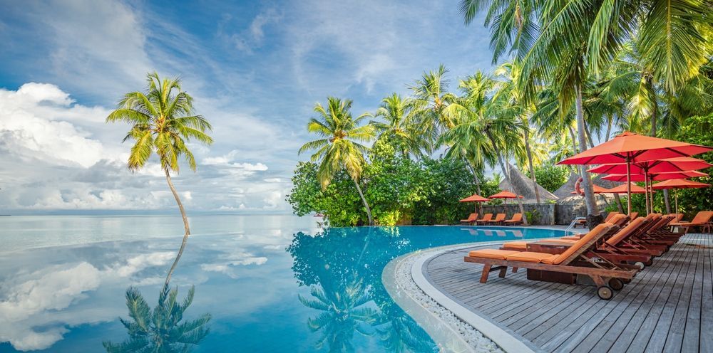 Tropical infinity pool with lounge chairs, red umbrellas, palm trees, and ocean view at sunset