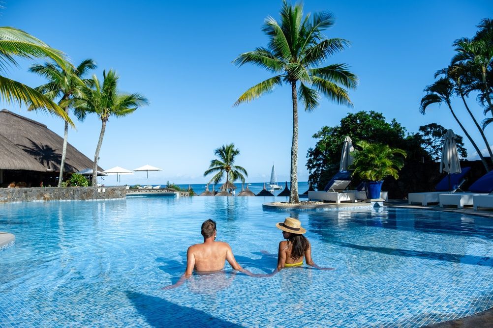 Two people relax in a blue infinity pool overlooking the ocean, with palm trees and a clear sky.