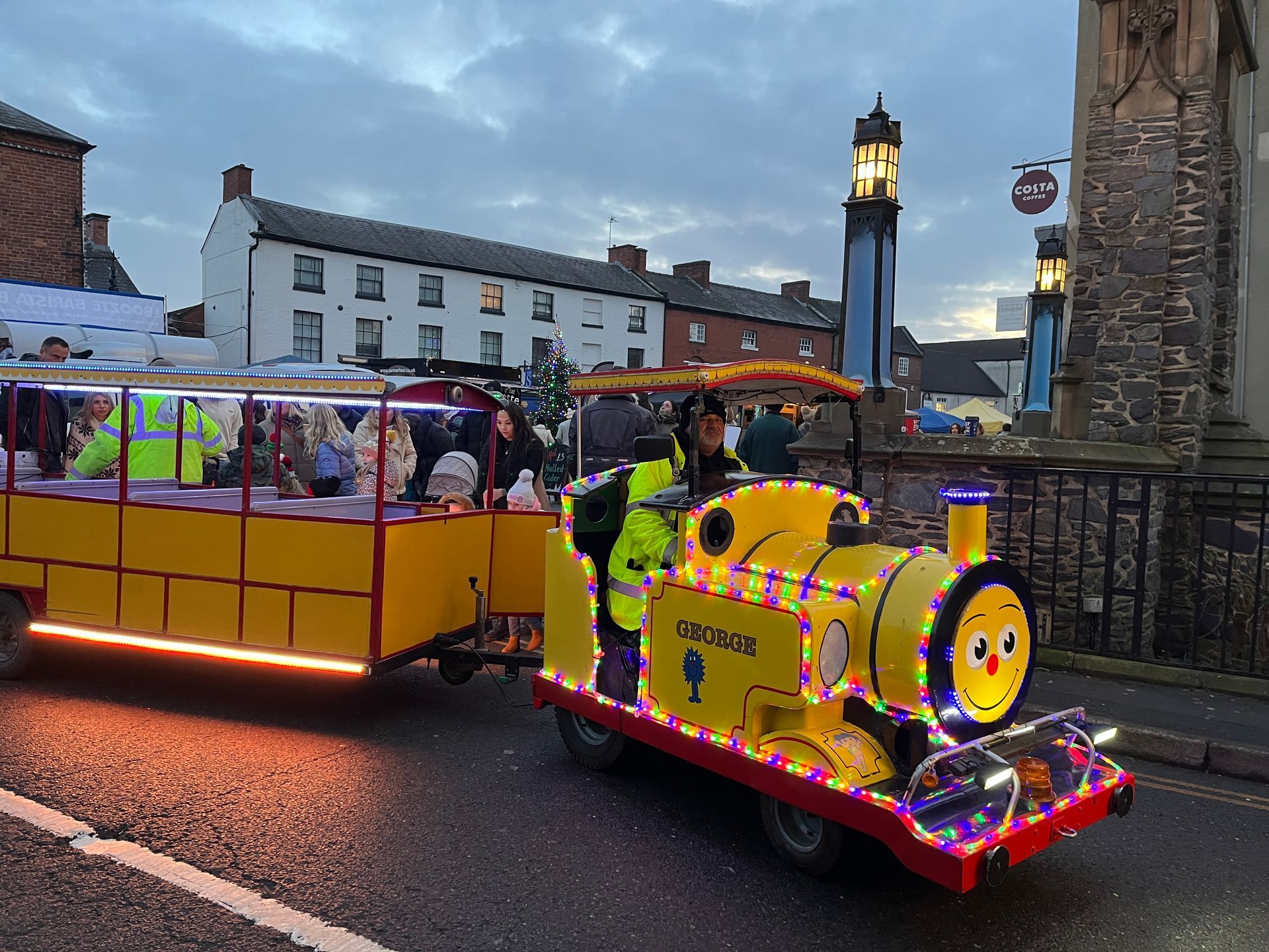 A yellow train with a trailer attached to it is driving down a street.