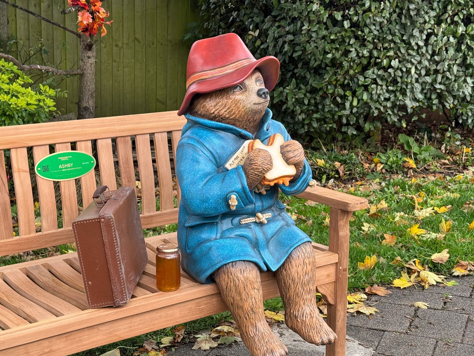 Paddington Bear statue on a bench, eating a sandwich, holding suitcase, jar of honey beside him.