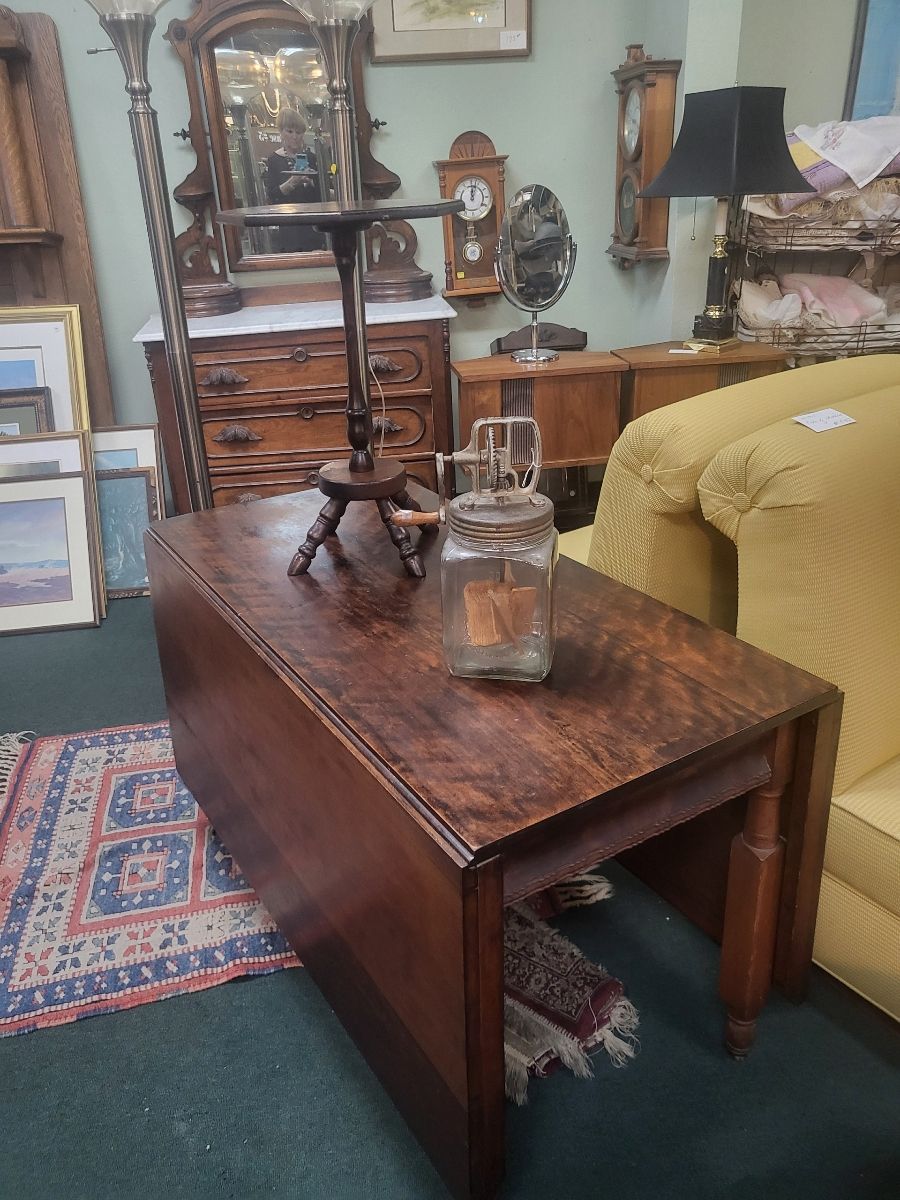 Antique table with glass jar and small stand in a shop; rug on the floor and other furniture in the background.