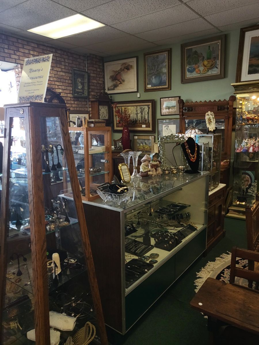 Antique shop interior with display cases of jewelry, paintings on the walls, and a small wooden chair.