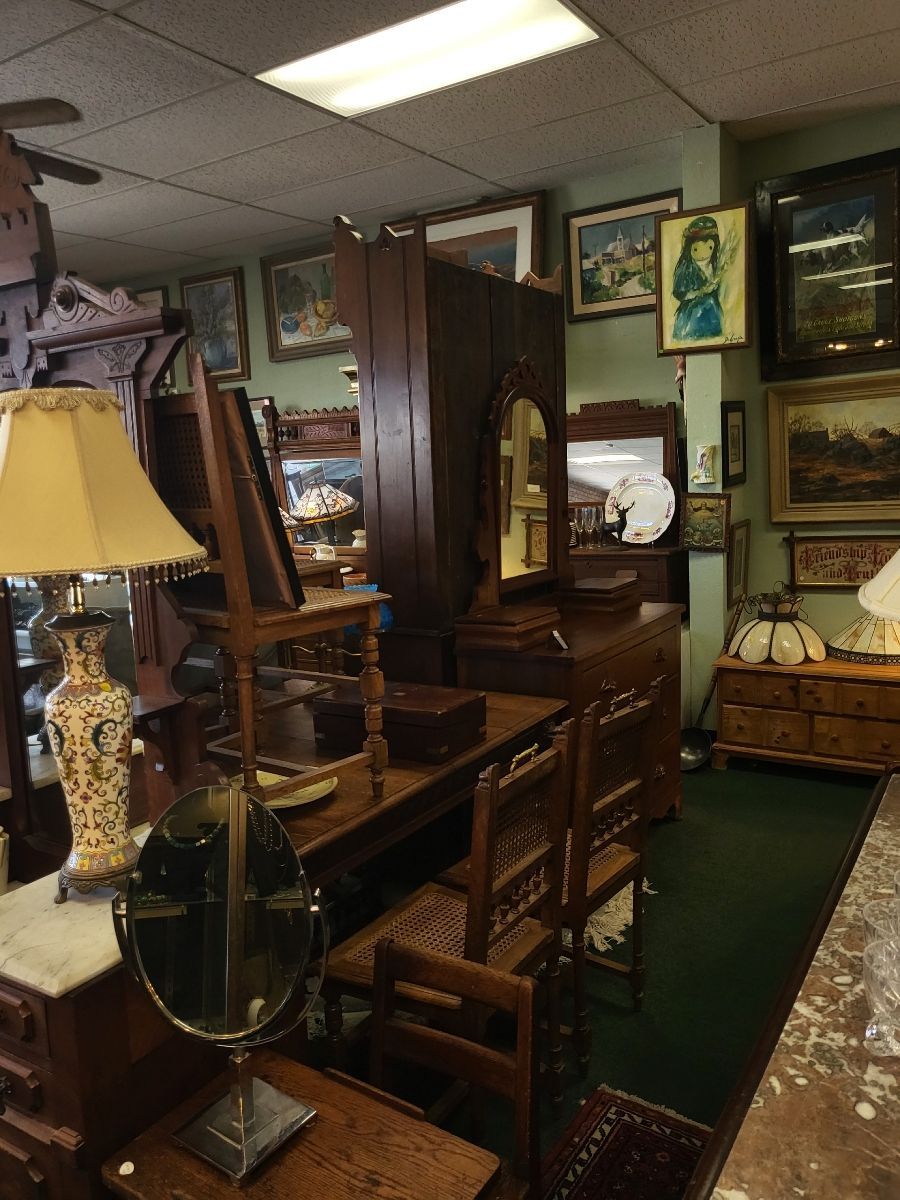 Antique furniture display in a shop. Dark wood dresser, chairs, and lamps in a room with paintings.