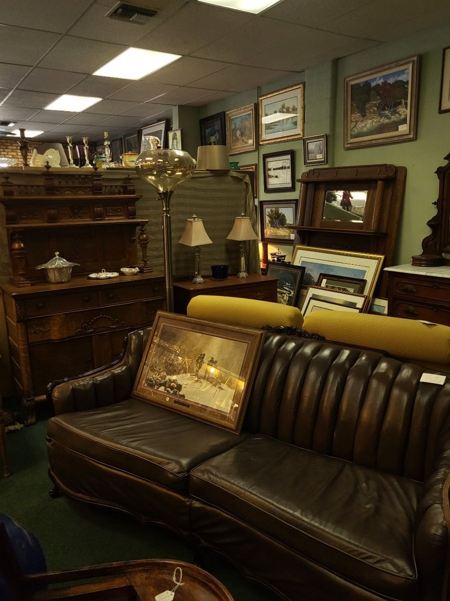 An antique shop filled with furniture, paintings, and lamps, featuring a leather sofa and wooden dresser.