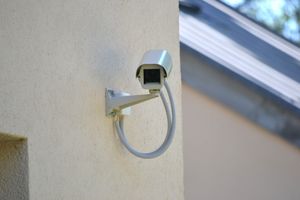 Telecamera di sicurezza montata su una parete beige con un cavo blu a forma di anello e il profilo del tetto nelle vicinanze.