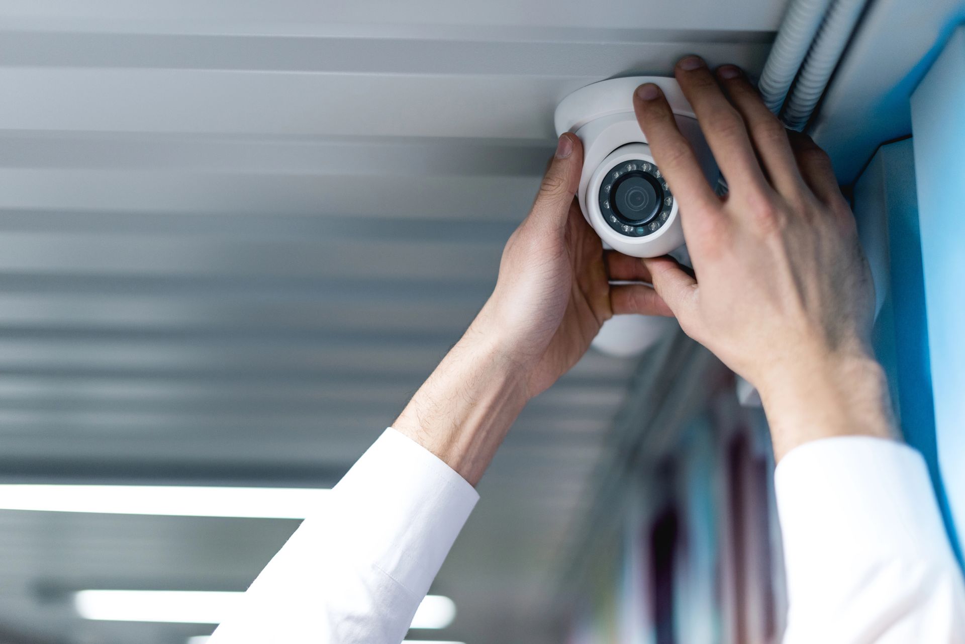 Mani che regolano una telecamera di sicurezza a soffitto in un ambiente interno, con le maniche bianche ben visibili.
