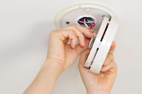 Mani che installano un rilevatore di fumo bianco sul soffitto, con i fili elettrici a vista.