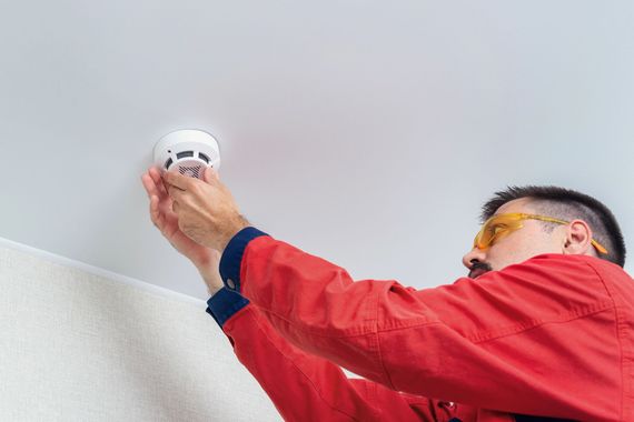 Tecnico in giacca rossa che installa un rilevatore di fumo a soffitto bianco con un attrezzo