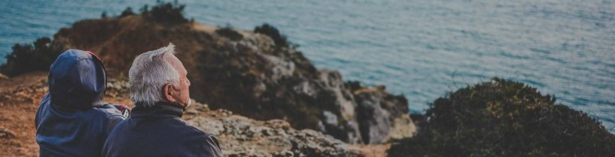 Two people in jackets sit on a rocky cliff, looking out over the ocean at sunset.
