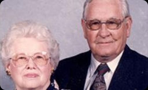 Portrait of an older couple in formal attire, both wearing glasses and facing the camera with neutral expressions