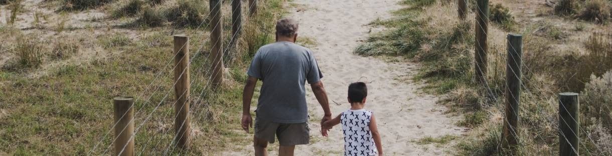 Two people walking hand in hand down a dirt path between wooden posts and dry grass