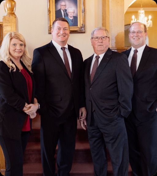 Four people in suits posing indoors near a staircase and chandelier