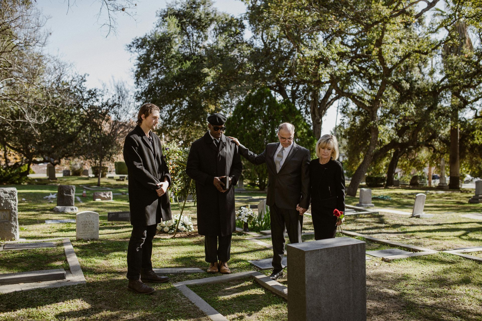 Four people stand in a cemetery beside a grave marker, appearing to mourn in a wooded, sunlit setting.