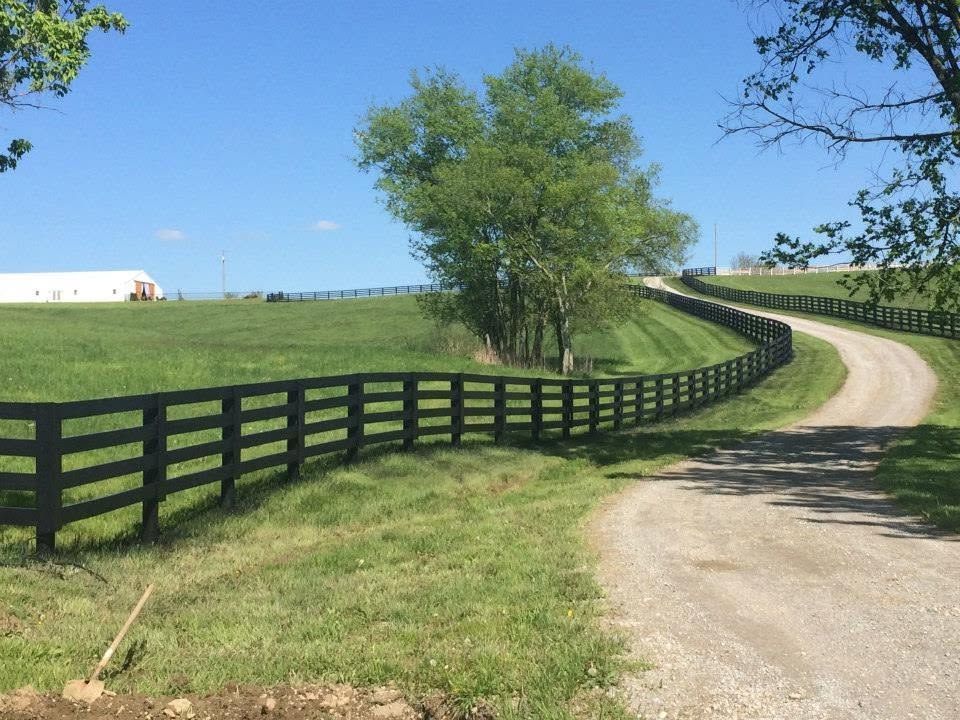 Curving gravel farm road beside black fence and green trees under a clear blue sky
