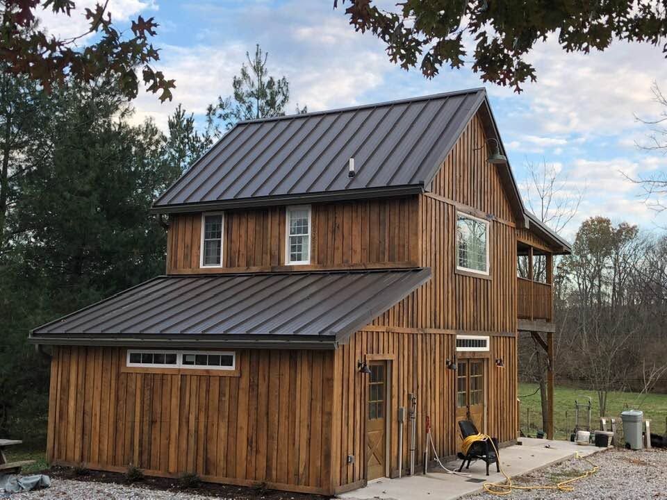 Small two-story wooden house with a dark metal roof, set in a rural yard with trees and gravel driveway.