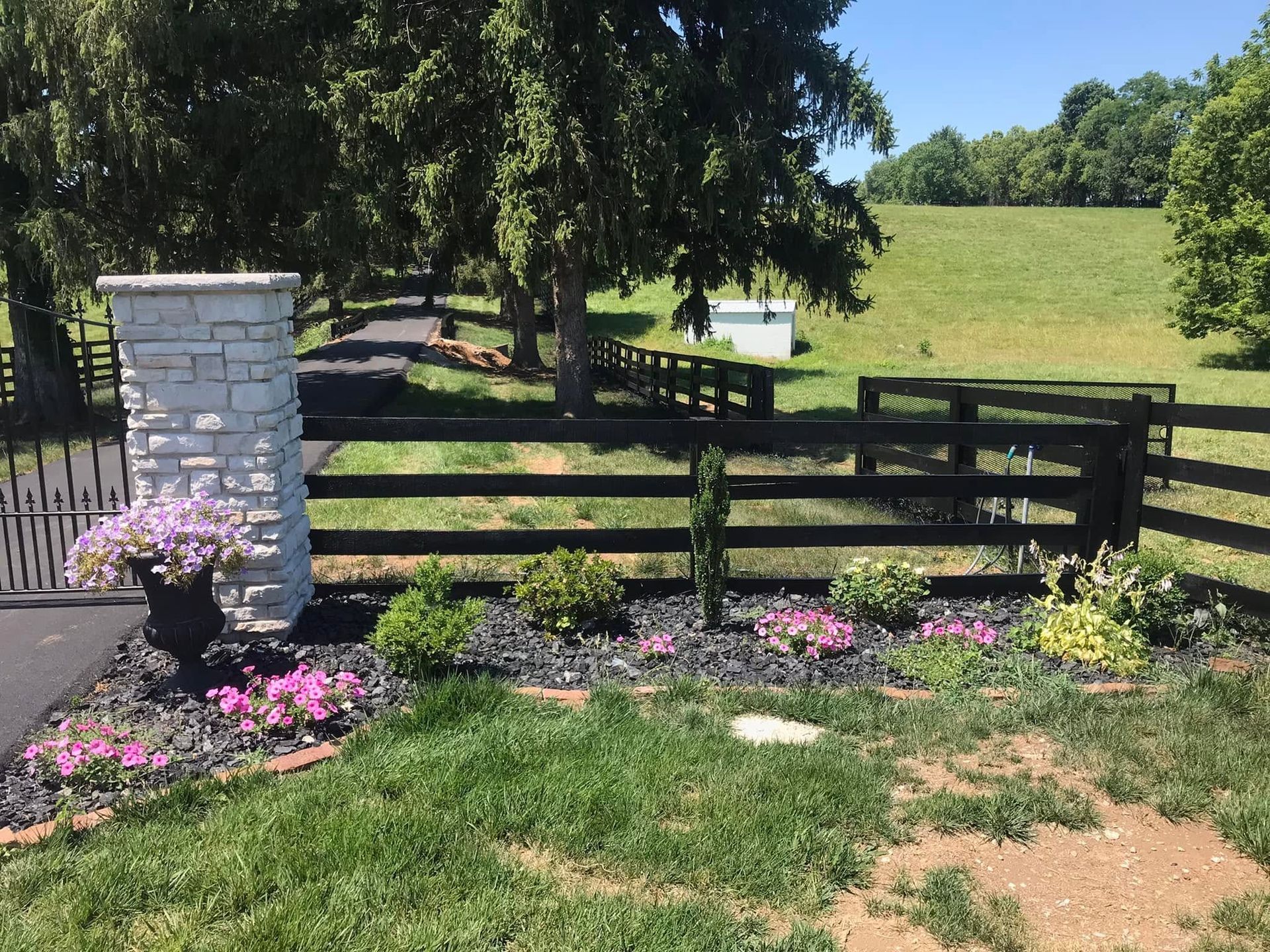Black wooden fence beside a white stone pillar, pink flowers, and a grassy field under sunny trees