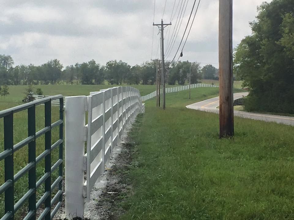 White fence beside a grassy roadside path with utility poles and a curved road in the distance